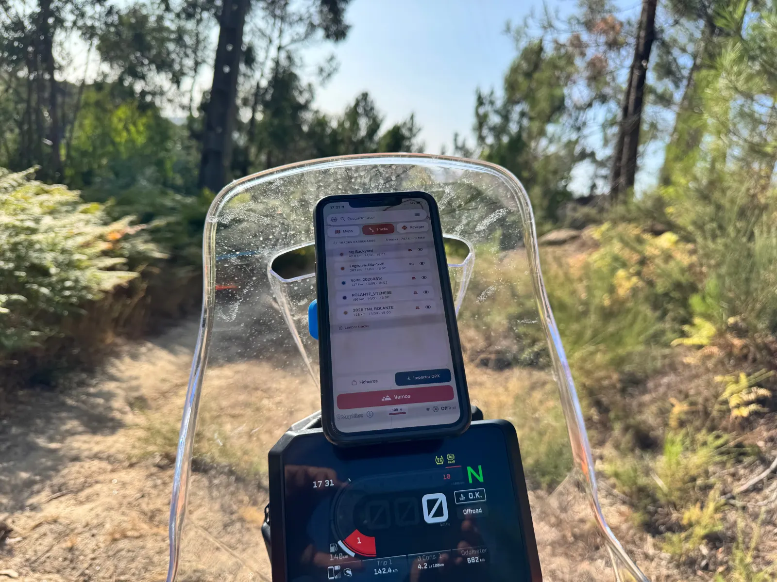 An iPhone running OffTrail mounted on an adventure motorcycle on a forest trail, with the bike's dashboard in off-road mode below.